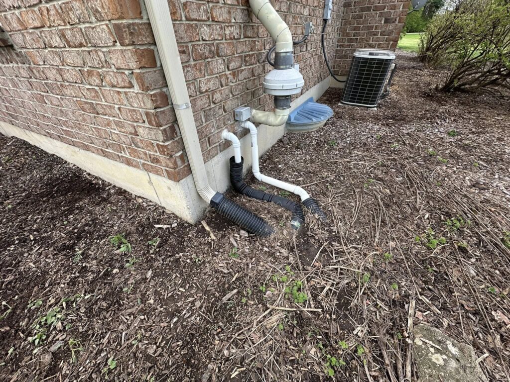 Before downspout drainage correction showing exposed pipes, unfinished ground, and poor water management near the foundation.